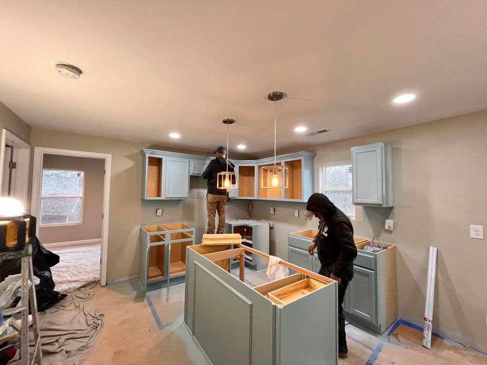 Workers installing light blue kitchen cabinets in a newly remodeled kitchen space.